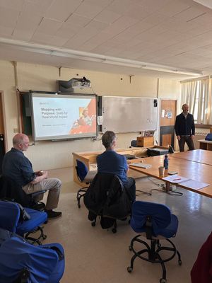 Img Description: Person standing at the front of a classroom with slides in the back