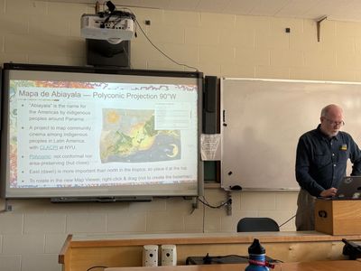 Img Description: Person standing at the front of a classroom with slides in the back